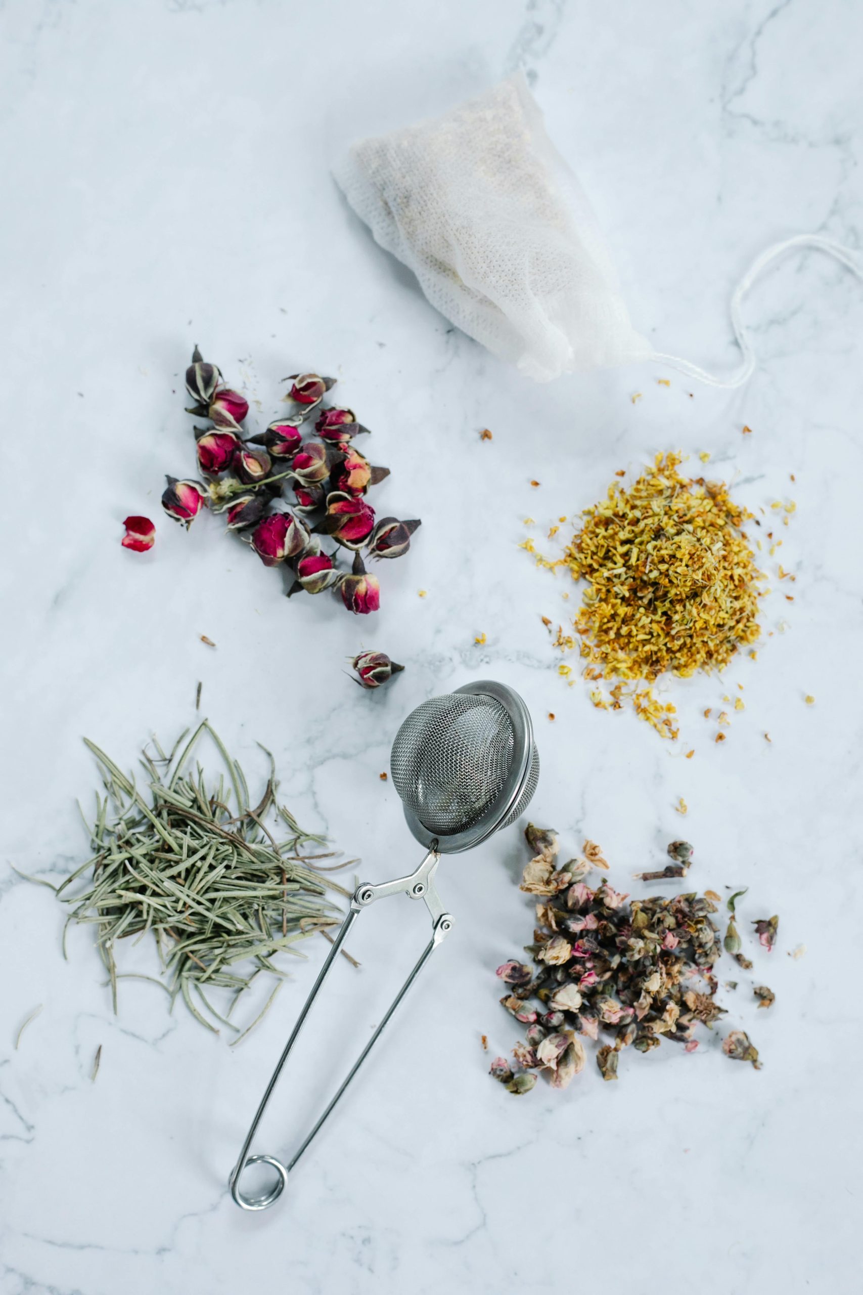 Top view of various dried herbal ingredients and tea infuser on a marble surface.